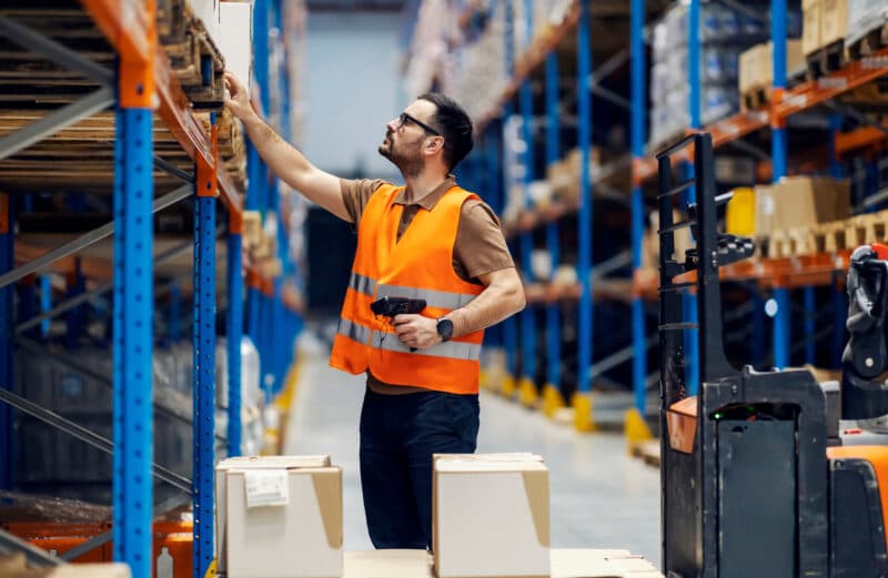 Warehouse worker in an orange safety vest reaches for a box while holding a barcode scanner.